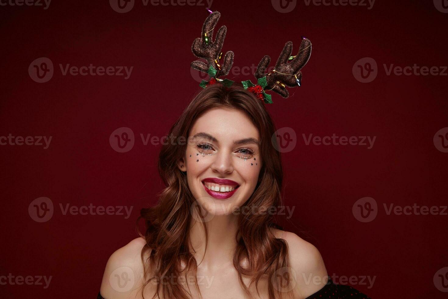 Close-up of cheerful young brown haired female with festive makeup wearing head horns while standing over claret background, smiling and expresses true positive emotions photo