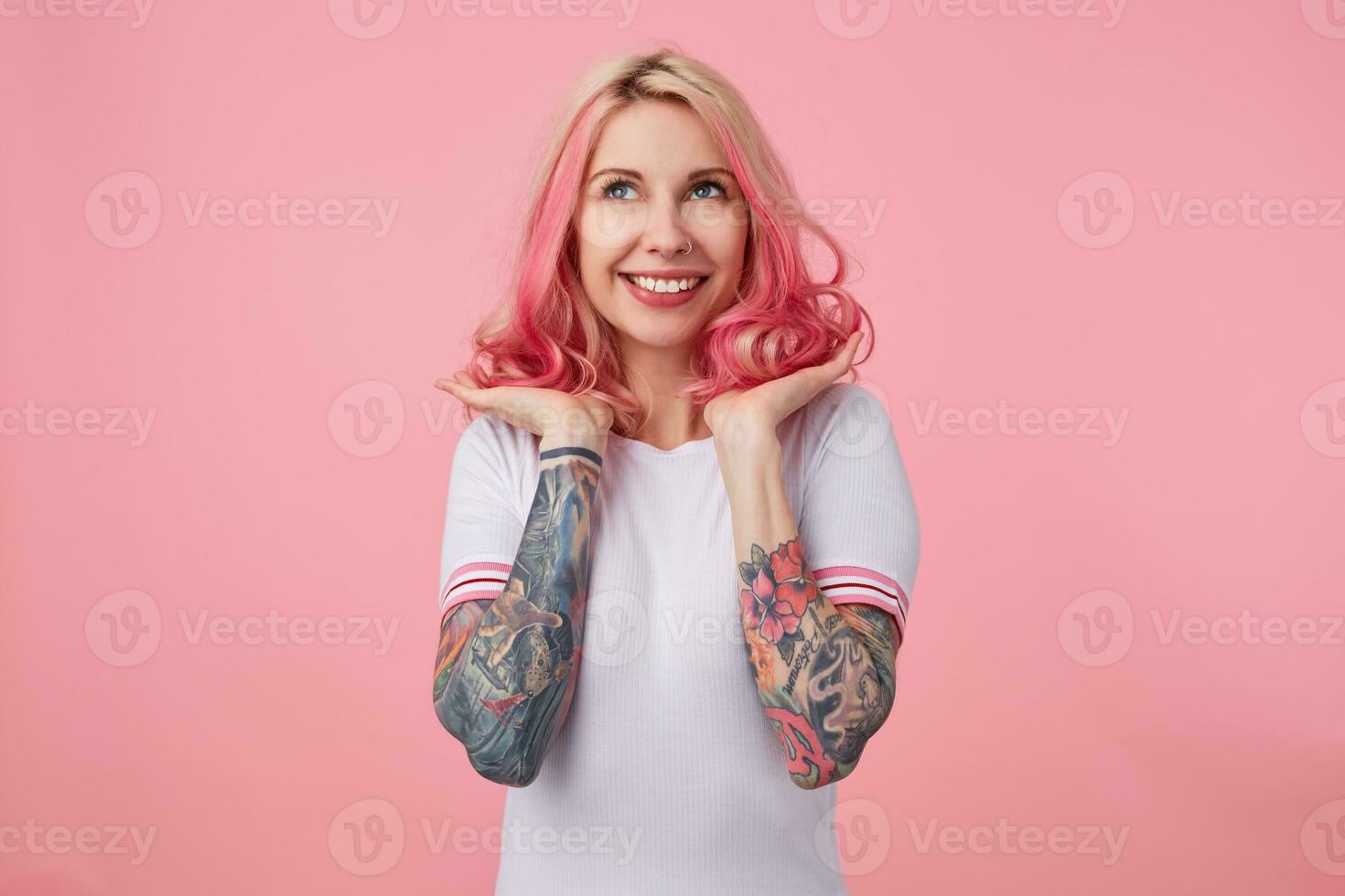 Portrait of young happy beautiful pink haired lady with tattooed hands, wears in white t-shirt, straightens her hair, happy with her new hair color and styling. Looks up, stands over pink background. photo