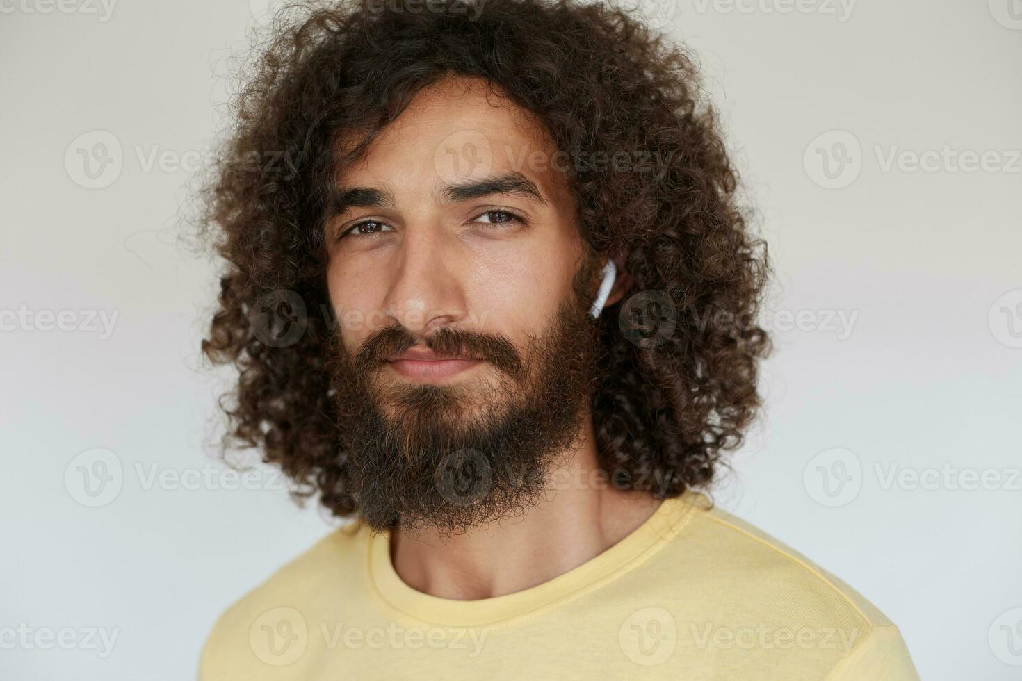 Close-up of beautiful young dark haired curly man with charming brown eyes looking positively to camera with light smile, wearing lush beard and listening to music in his headphones photo