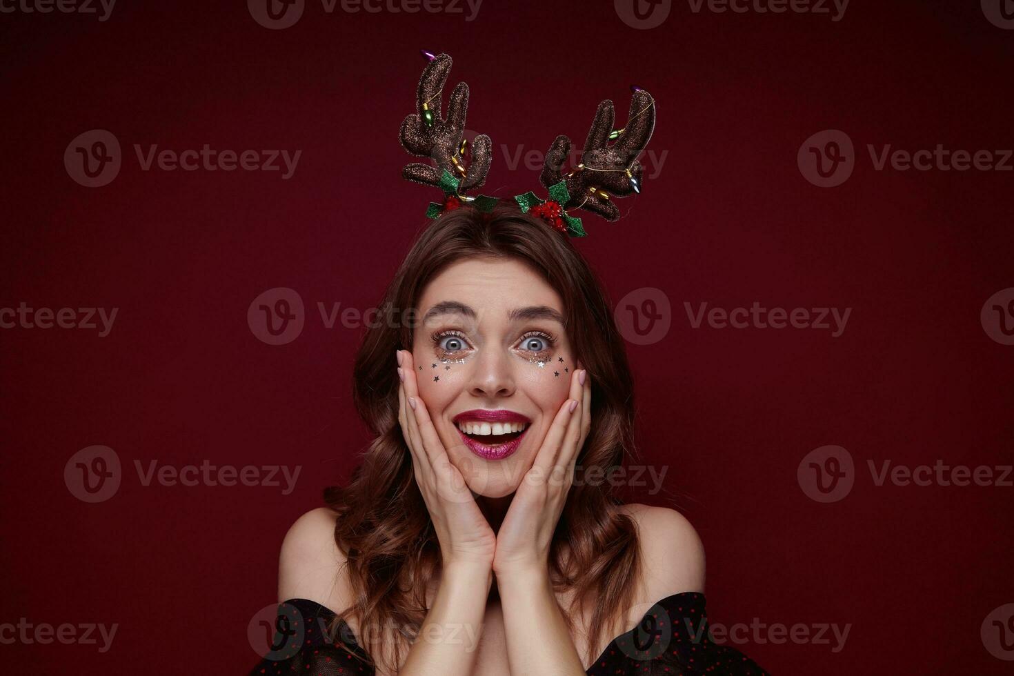 Surprised blue-eyed young brown haired woman with evening makeup wearing holiday hoop and looking at camera with wide eyes opened, keeping palms on cheeks and smiling widely photo