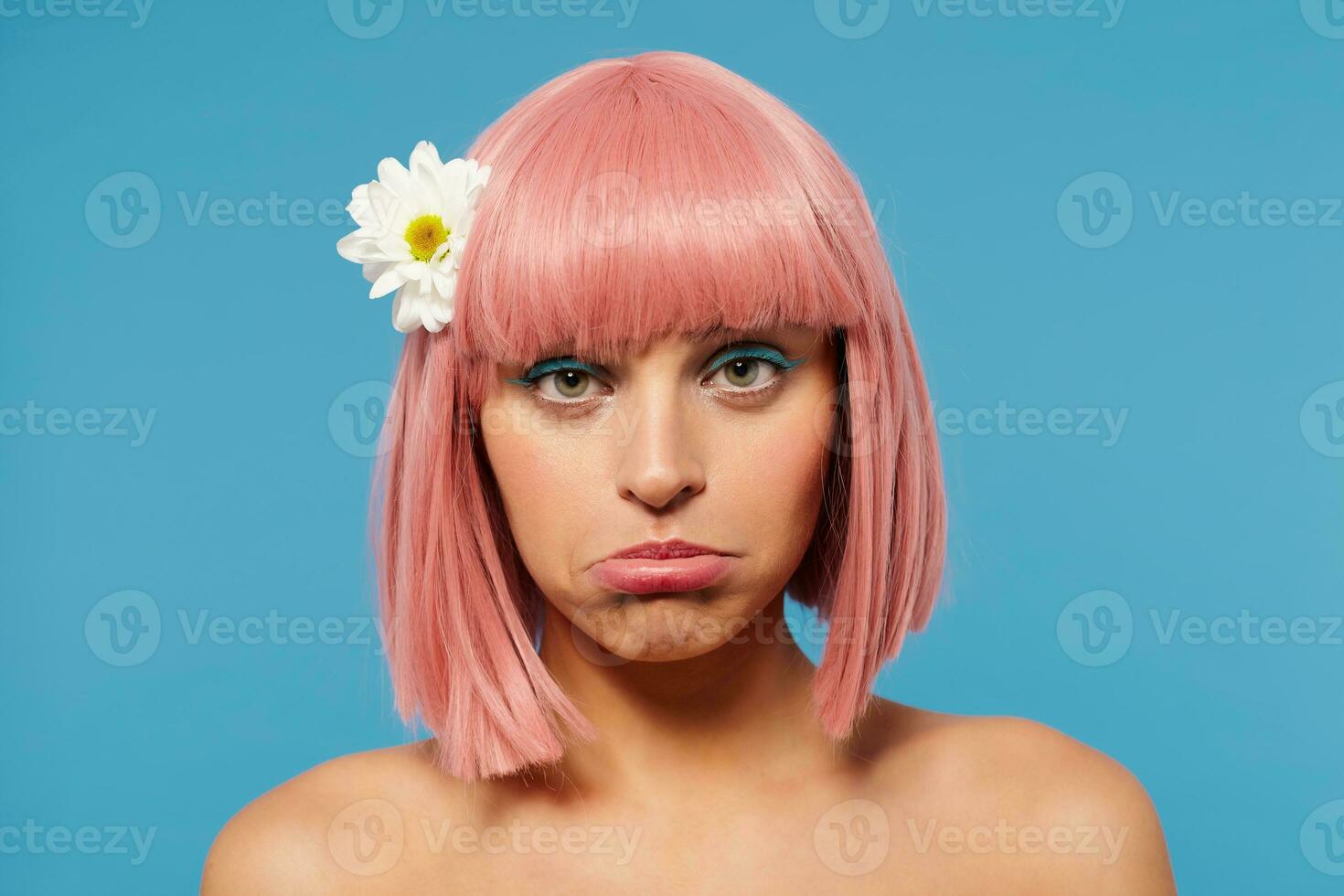 Close-up of young offended green-eyed lady with short pink hair wearing festive makeup while standing over blue background, twisting her mouth while looking sadly at camera photo