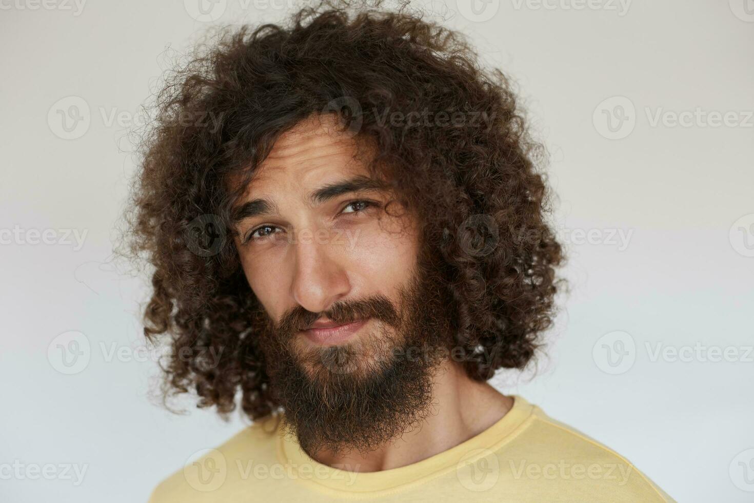 Studio photo of flirty young brunette curly man with beard looking playfully at camera with folded lips, squinting and wrinkling forehead against white background, dressed in casual clothes