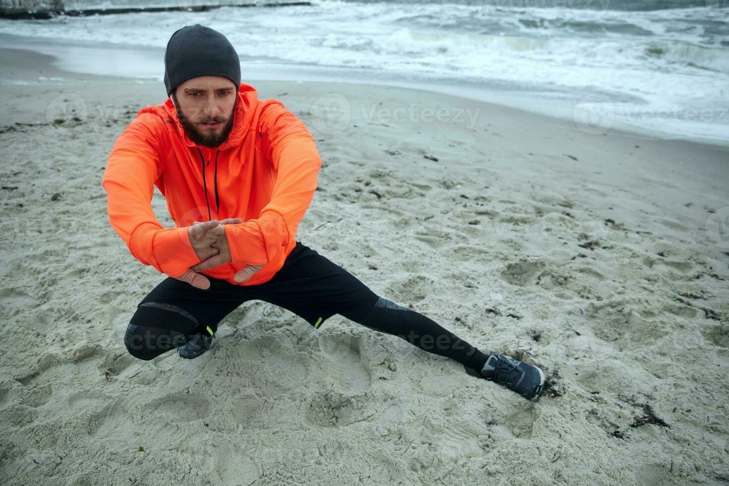 Outdoor photo of serious looking young bearded sportsman doing stretching exercise and keeping hands raised with crossed fingers, wearing athletic clothes and warm orange coat