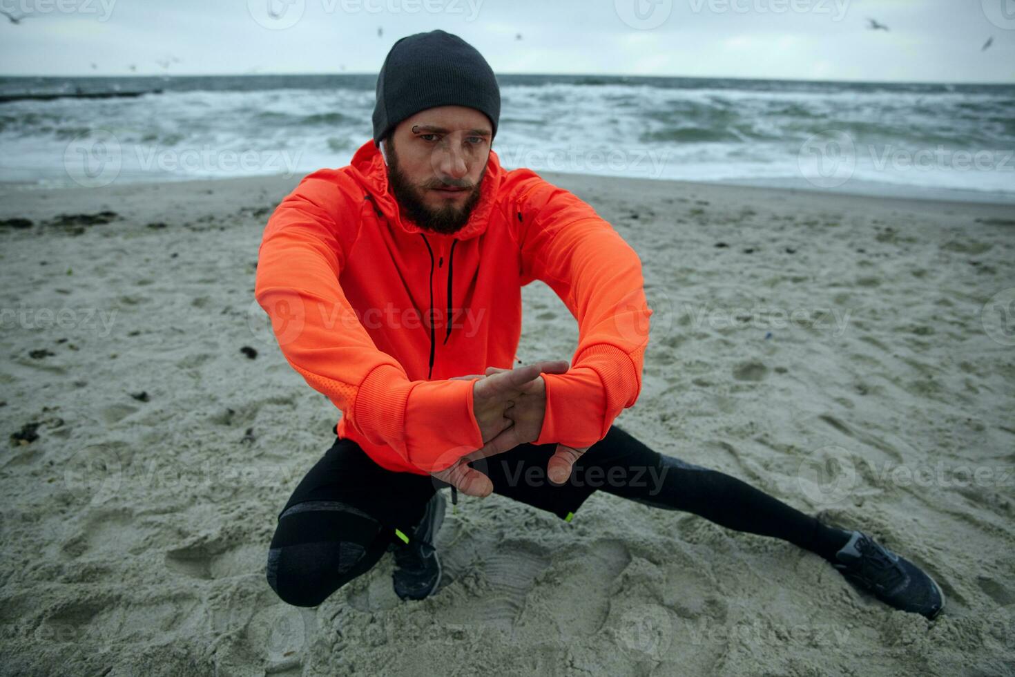 imagen de joven barbado masculino con ceja perforación mirando a cámara con grave cara mientras extensión su músculos antes de corriendo sesión, posando terminado playa ver foto