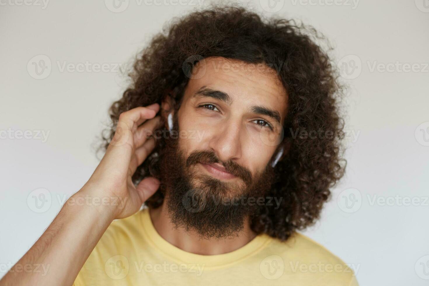Portrait of attractive brown-eyed young brunette man with curly hair and lush beard keeping hand behind his ear while looking at camera, wearing yellow t-shirt and headphones against white background photo