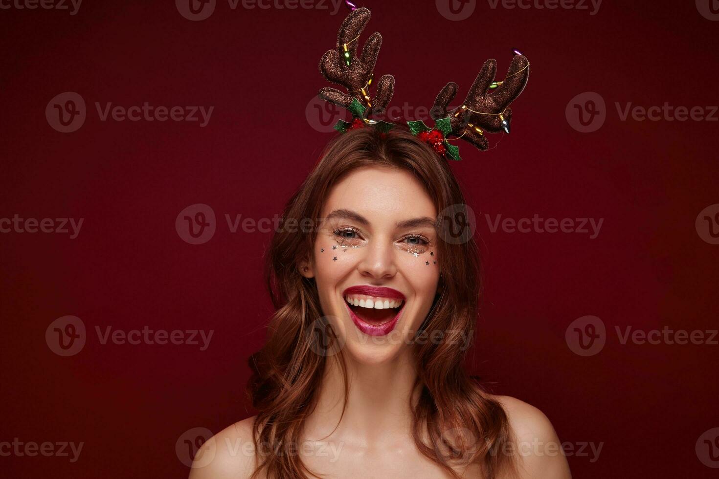 Close-up of joyful attractive young brunette lady with wavy hairstyle wearing holiday horns on christmas party, rejoicing while standing over claret background, expressing true positive emotions photo