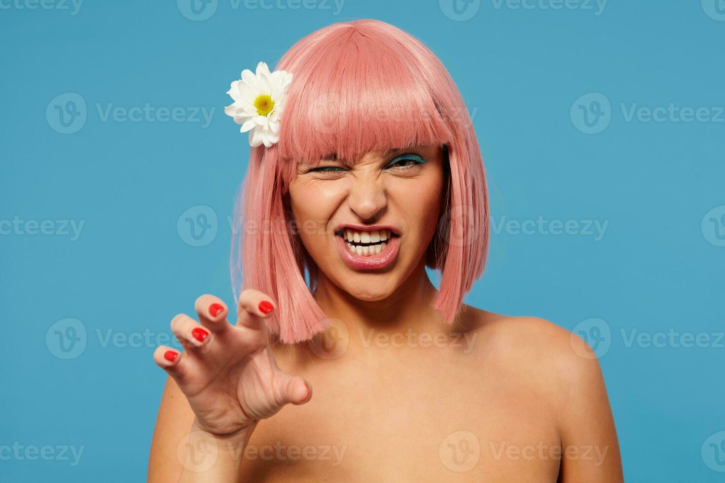 Playful young pretty lady wearing chamomile in her short pink hair while posing over blue background, showing her teeth and giving wink at camera while imitating cat's paw with raised hand photo