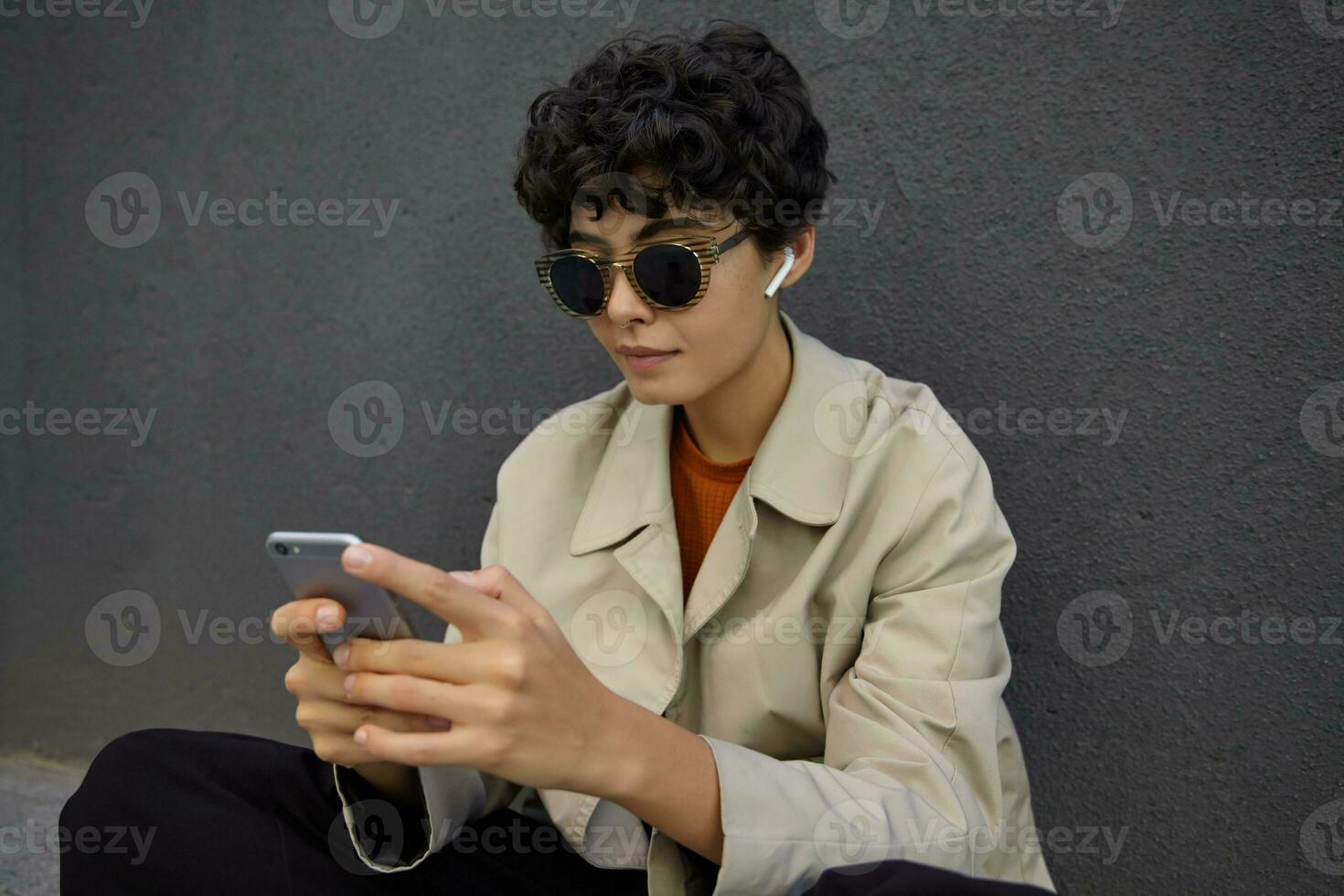 Portrait of stylish pretty brunette female with curly short hair wearing sunglasses and earphones while posing over concrete black wall,keeping smartphone in hands and looking at screen with calm face photo