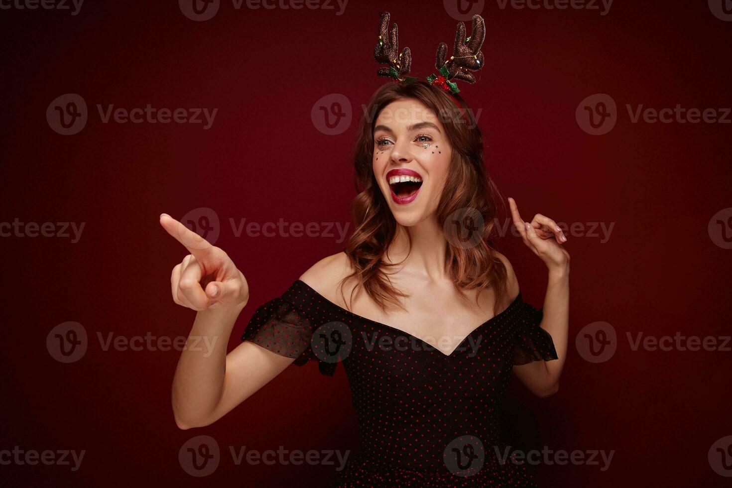 Christmasnew year themed portrait of young lovely brown haired woman in black elegant dress with red dots pointing cheerfully aside with forefinger and smiling widely, isolated over claret background photo