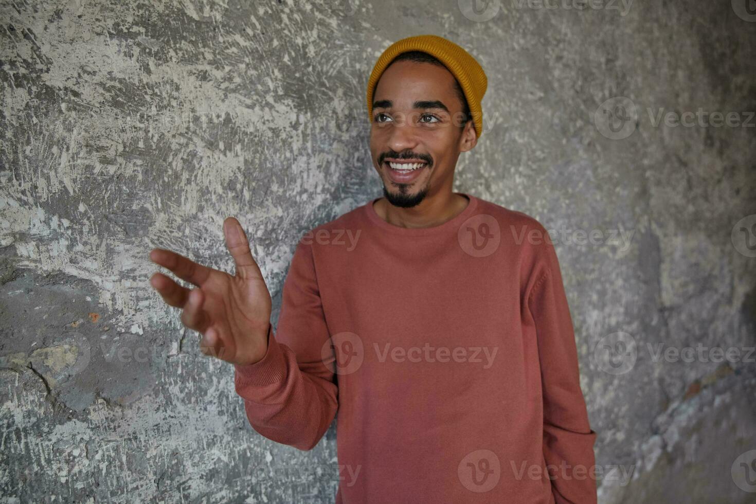 Indoor shot of joyful lovely bearded dark skinned man smiling sincerely with raised hand, looking aside while posing over concrete background, showing his pleasant emotions photo