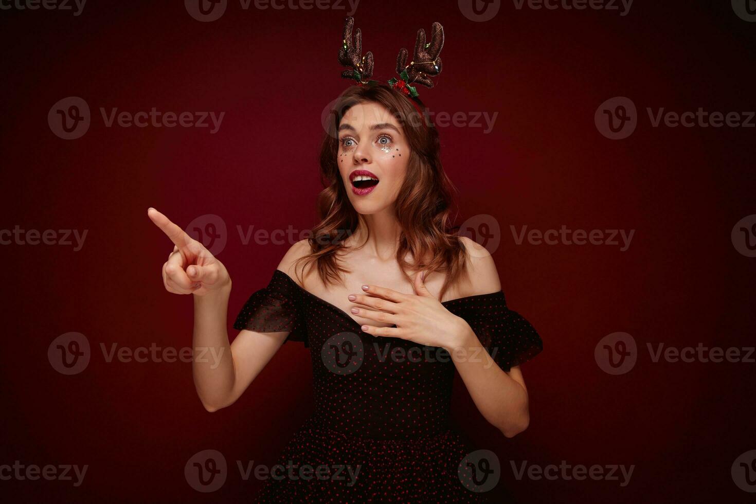 Open-eyed young beautiful brunette lady with festive hairstyle dressed in black girlish dress and christmas hoop showing aside amazedly with wide mouth opened, isolated over claret background photo
