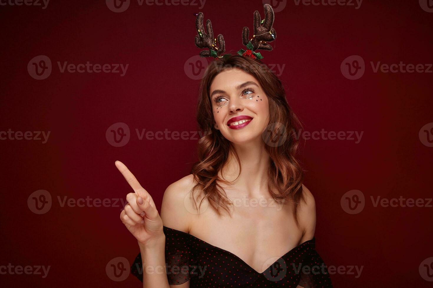 Portrait of young positive pretty brown haired female with wavy hairstyle looking happily aside and smiling sincerely, preparing for new year party and wearing holiday horns over claret background photo