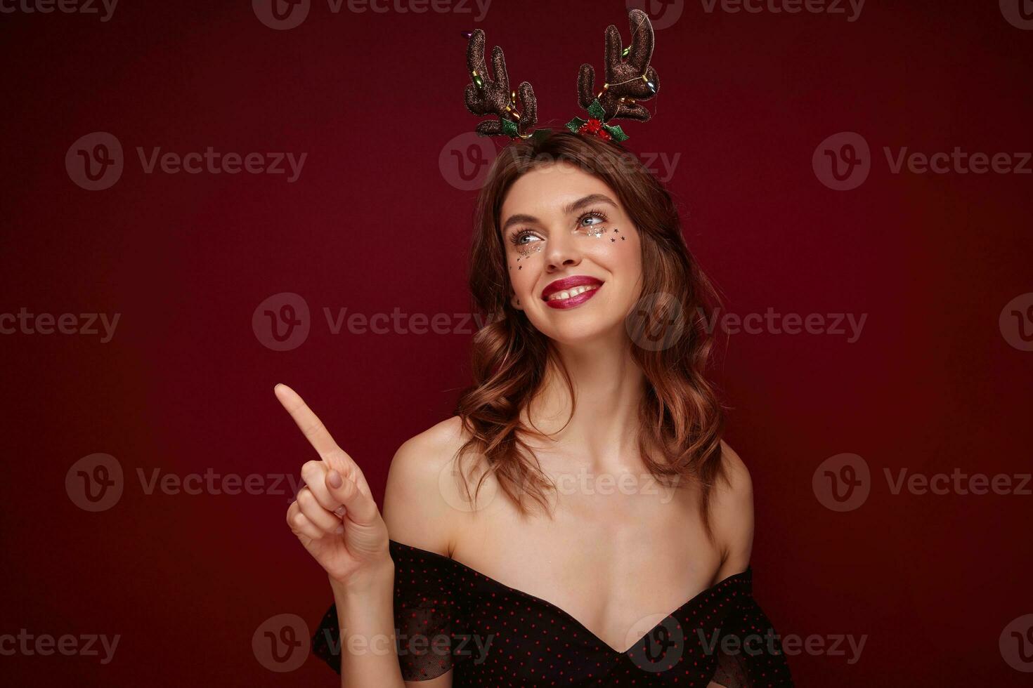 retrato de joven positivo bonito marrón peludo hembra con ondulado peinado mirando felizmente aparte y sonriente atentamente, preparando para nuevo año fiesta y vistiendo fiesta cuernos terminado burdeos antecedentes foto