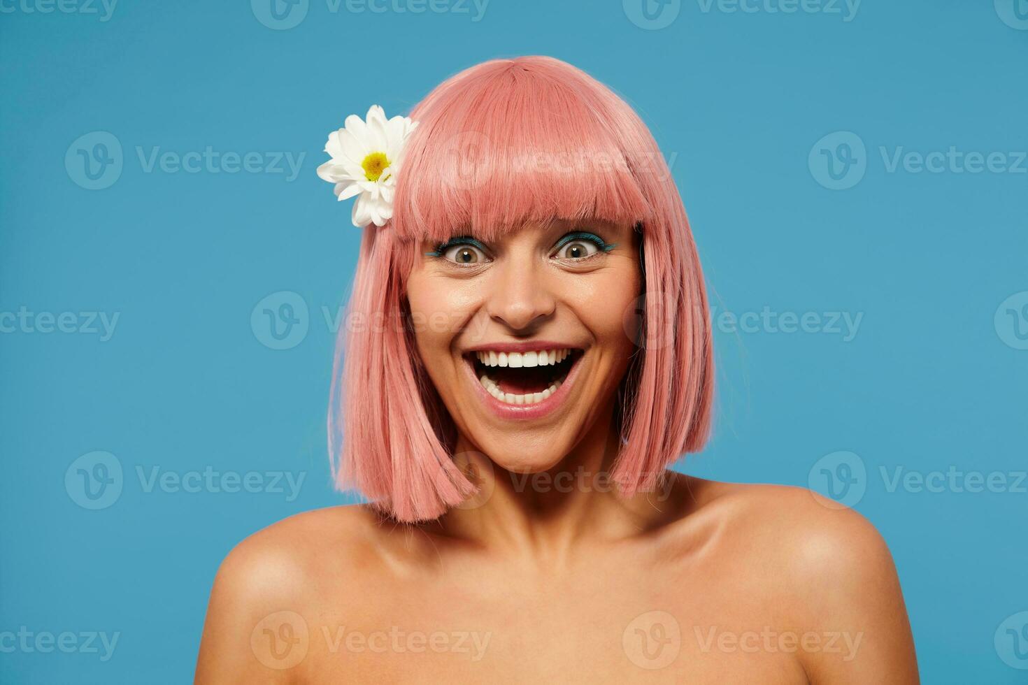 Excited young attractive pink haired female looking happily at camera with wide eyes and mouth opened, showing her pleasant emotions while posing over blue background photo