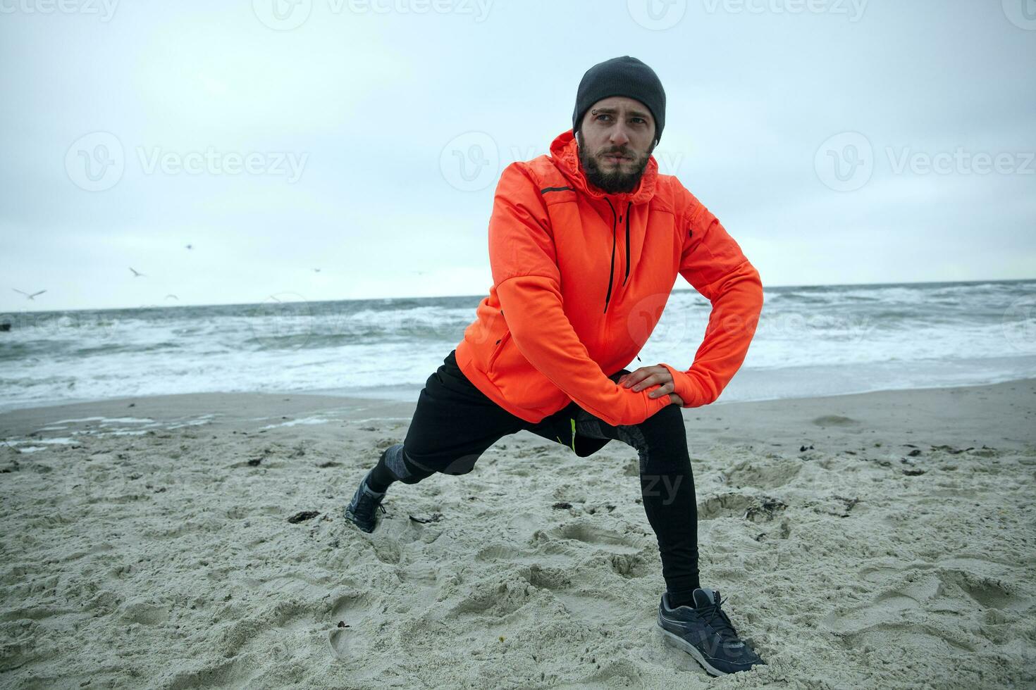 Horizontal shot of attractive young bearded male stretching his muscles before running exercise, standing over coastline of sea on gray cold weather, goes in for sport every morning photo