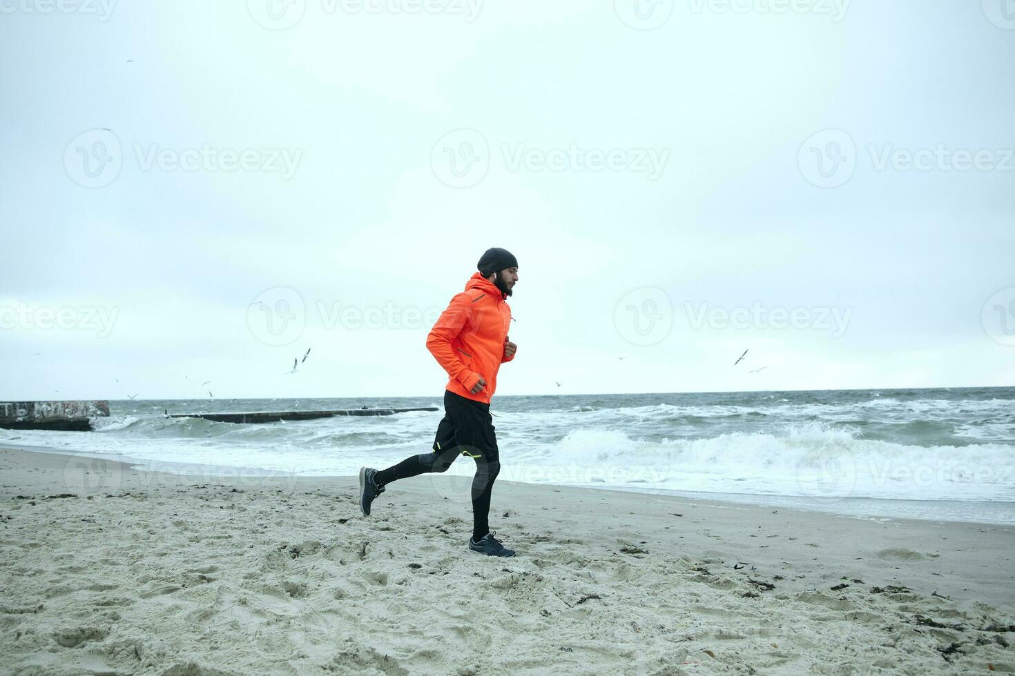 longitud total foto de joven barbado deportista vestido en atlético ropa y calentar naranja Saco trabajando fuera y corriendo por playa en gris frío clima. aptitud masculino modelo