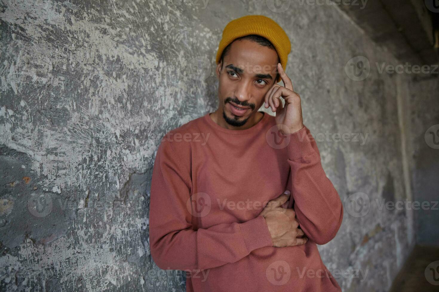 Serious looking pretty dark skinned guy with beard wearing pink sweater and mustard headdress, leaning on concrete wall while having grave conversation photo