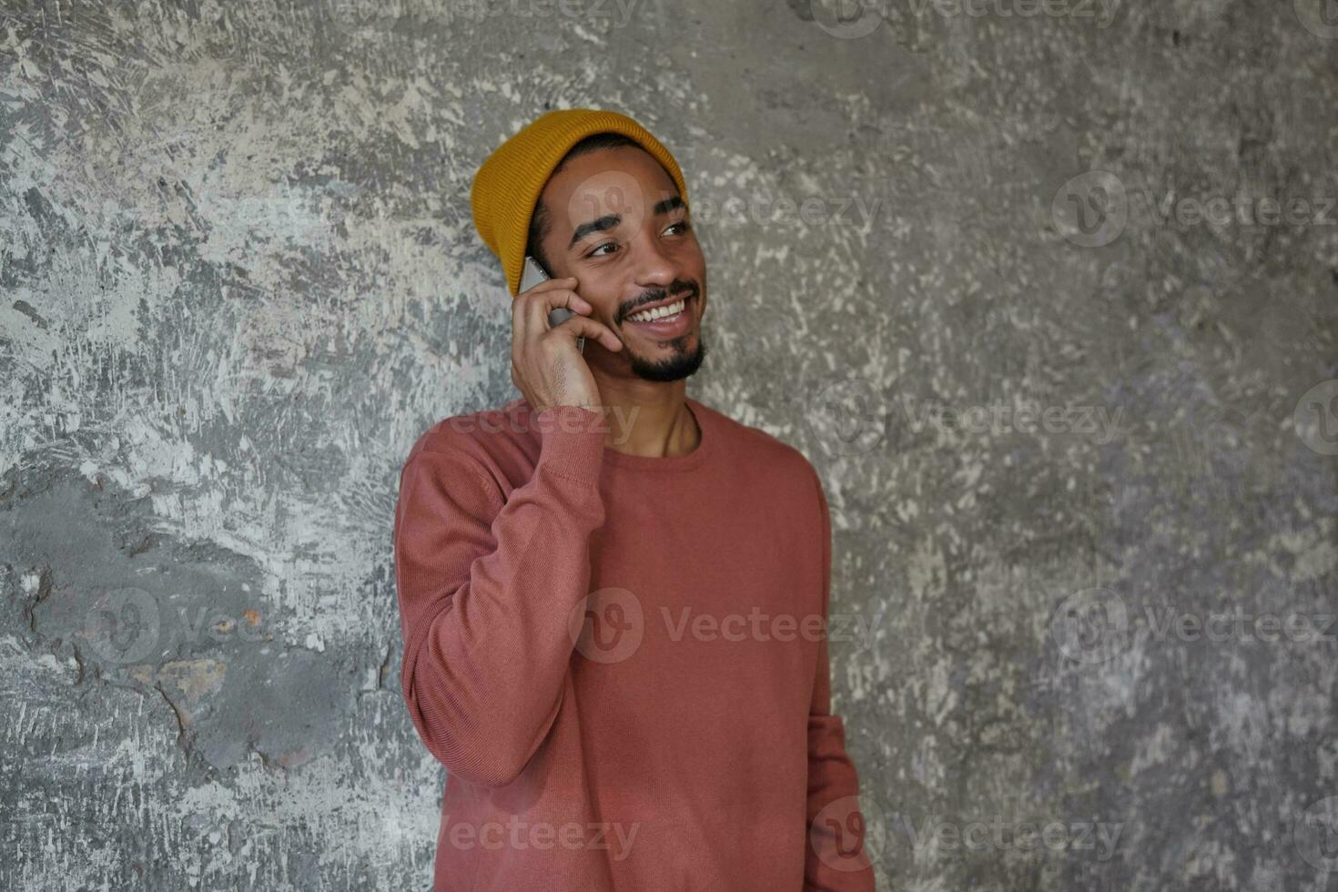 Cheerful bearded dark skinned male with charming brown eyes looking aside and smiling happily while having nice talk on phone, posing over concrete background in casual clothes photo