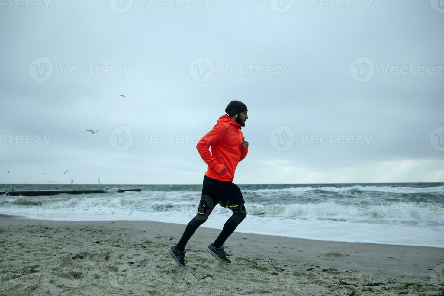 Outdoor shot of young bearded runner with headphones jogging on sandy beach before working day, leads healthy active lifestyle, being calm and concentrated photo