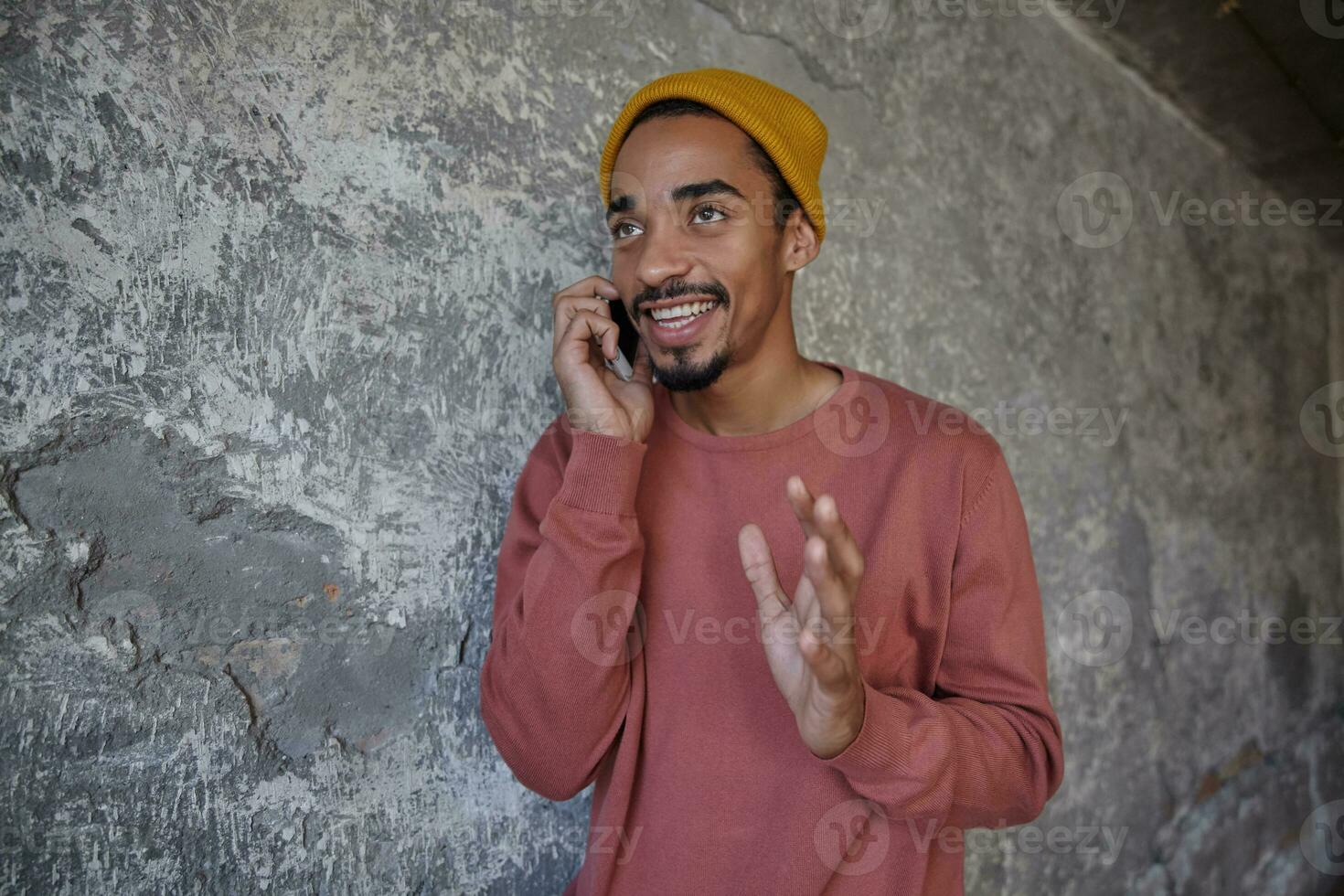 Shot of positive attractive young bearded man with dark skin raising hand while talking on phone, sharing news with his friend and having nice day, standing over concrete wall photo