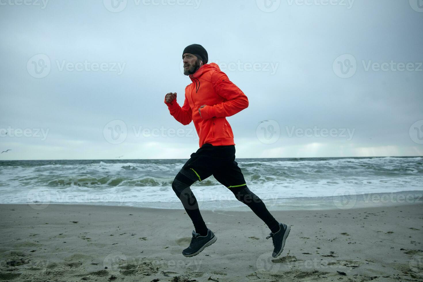 Side view of young sporty brunette bearded man in warm orange coat and black athletic clothes exercising outdoors before working day, goes in for sport every morning. Fitness and sport concept photo