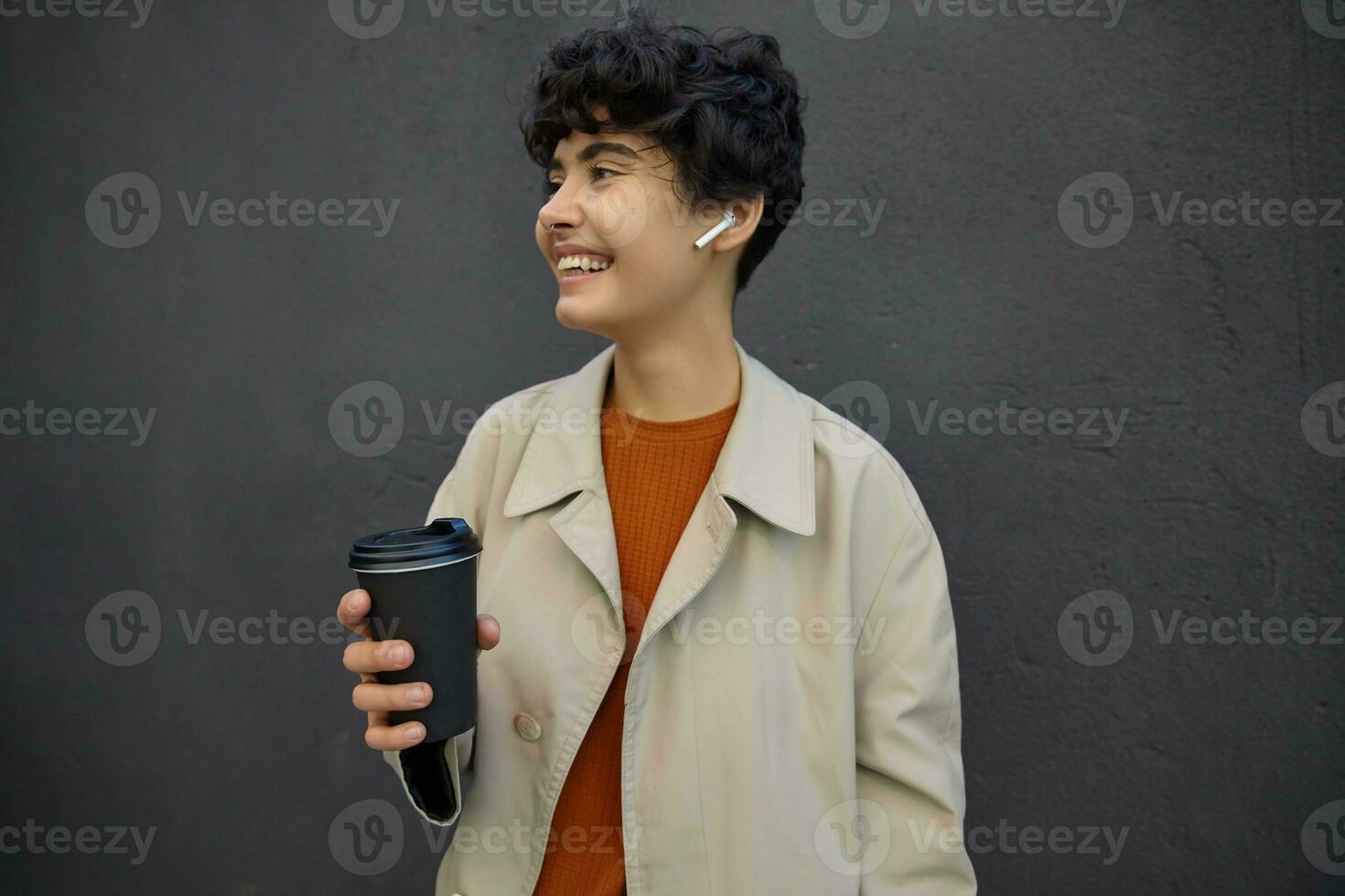 Positive young attractive brunette woman with short haircut keeping takeaway coffee and looking aside cheerfully with joyful smile, wearing trendy outfit while posing over urban environment photo