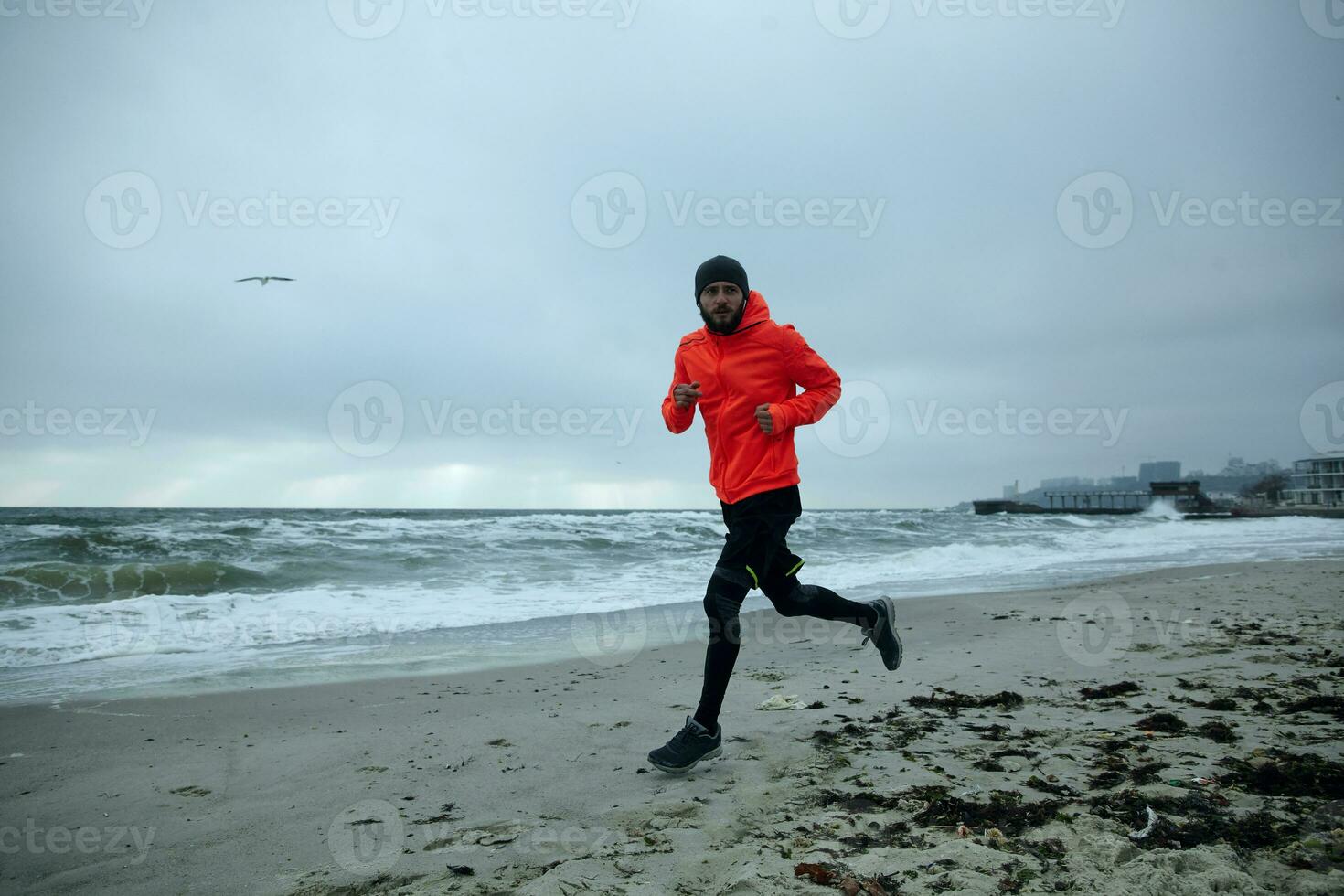 longitud total imagen de joven activo barbado masculino vestido en calentar deportivo ropa trabajando fuera y corriendo por playa en frío melancólico clima. deporte y sano estilo de vida concepto foto