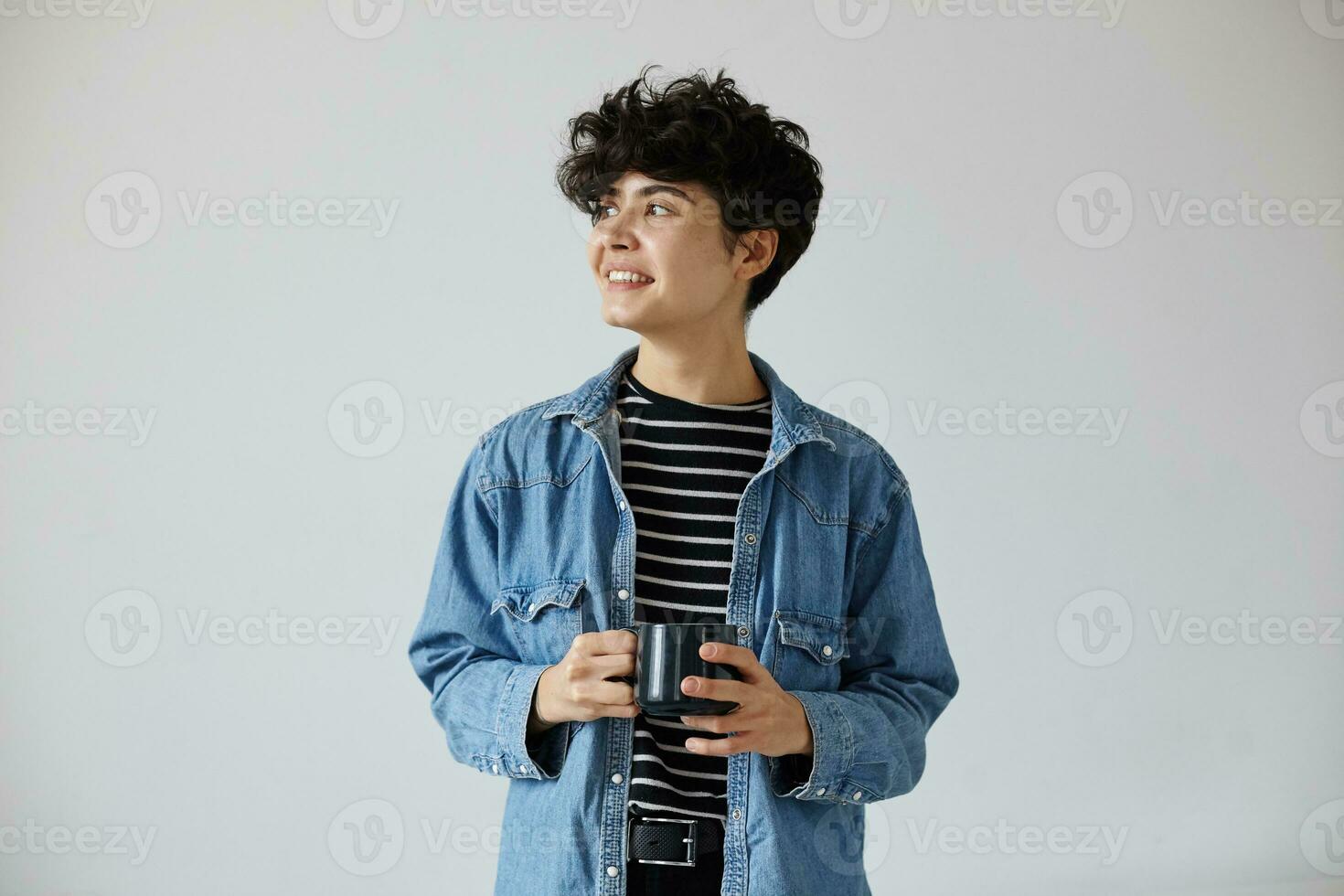 Stylish young pretty short haired curly brunette female holding cup of tea in raised hands and looking positively aside with pleasant smile, standing against white background photo