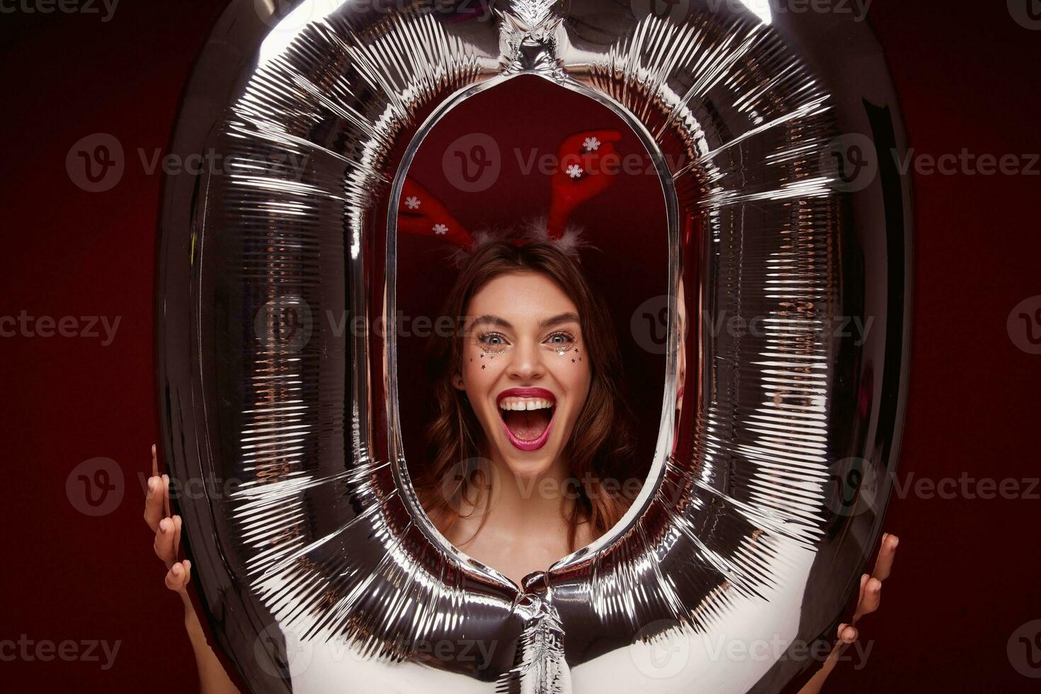 Indoor photo of overjoyed young brown haired female wearing festive makeup while posing over big air balloon, rejoicing nice new year party together with friends, isolated over claret background