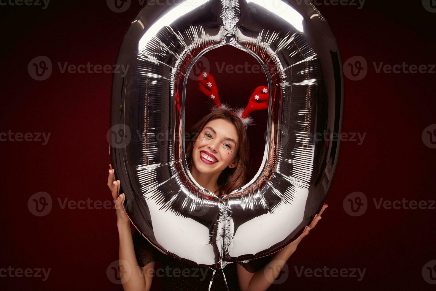 Studio shot of joyful beautiful young brunette female lady with evening makeup posing over claret background with air number balloon, preparing for new year party photo