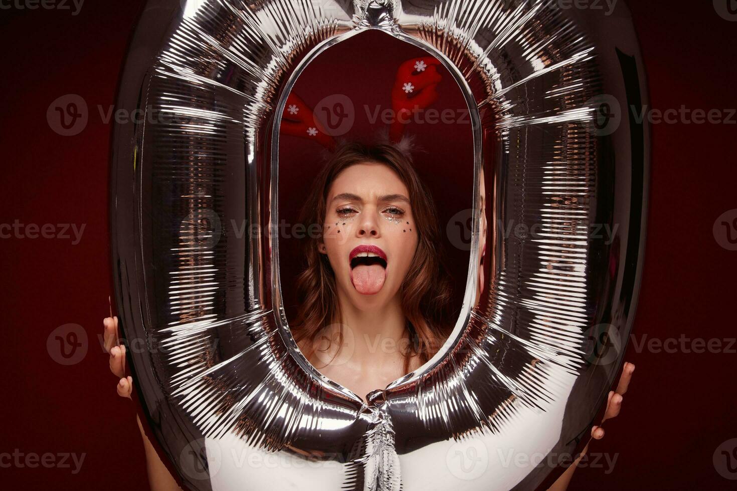 Close-up of young funny pretty female with wavy hairstyle fooling and making faces while posing over claret background with huge air number balloon. Holiday preparation and celebration concept photo