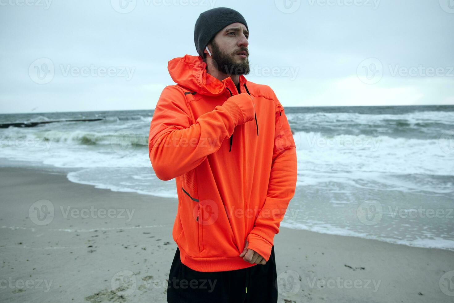 Serious young brunette bearded sportsman frowning eyebrows and looking ahead while zipping up his athletic orange coat, standing over seaside on cold overcast weather photo