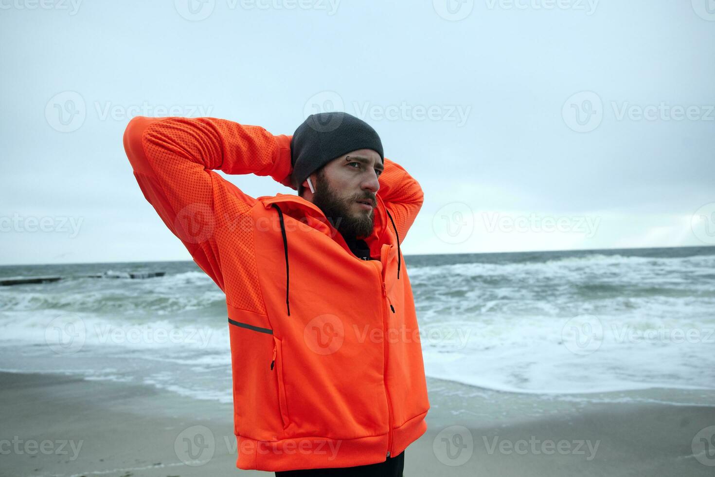 Portrait of young dark haired bearded male with eyebrow piercing dressed in warm sporty clothes folding hands behind his head and looking thoughtfully aside photo
