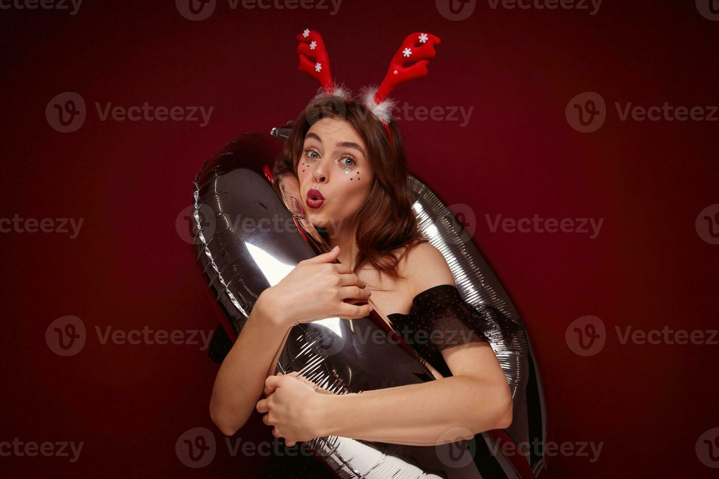 Funny shot of young pretty brunette lady wearing festive makeup while posing over claret background, enjoying x-mas theme party and posing with helium balloon photo