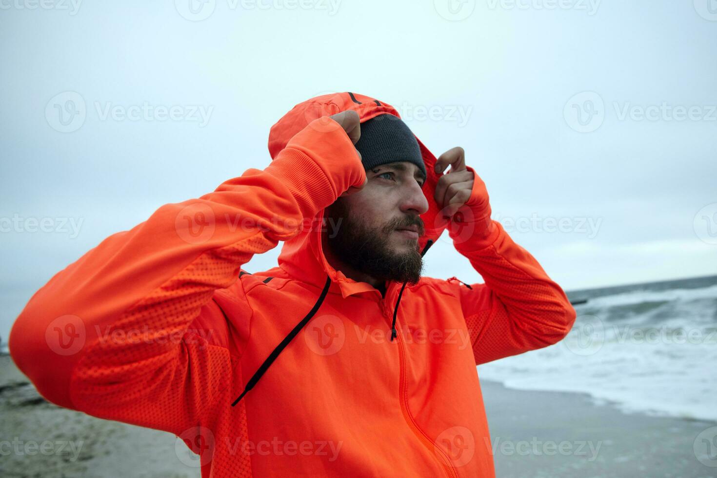 Close-up of young handsome bearded man holding raised hand on hood of his warm athletic orange coat while breathing fresh air over seaside, looking ahead with calm face photo