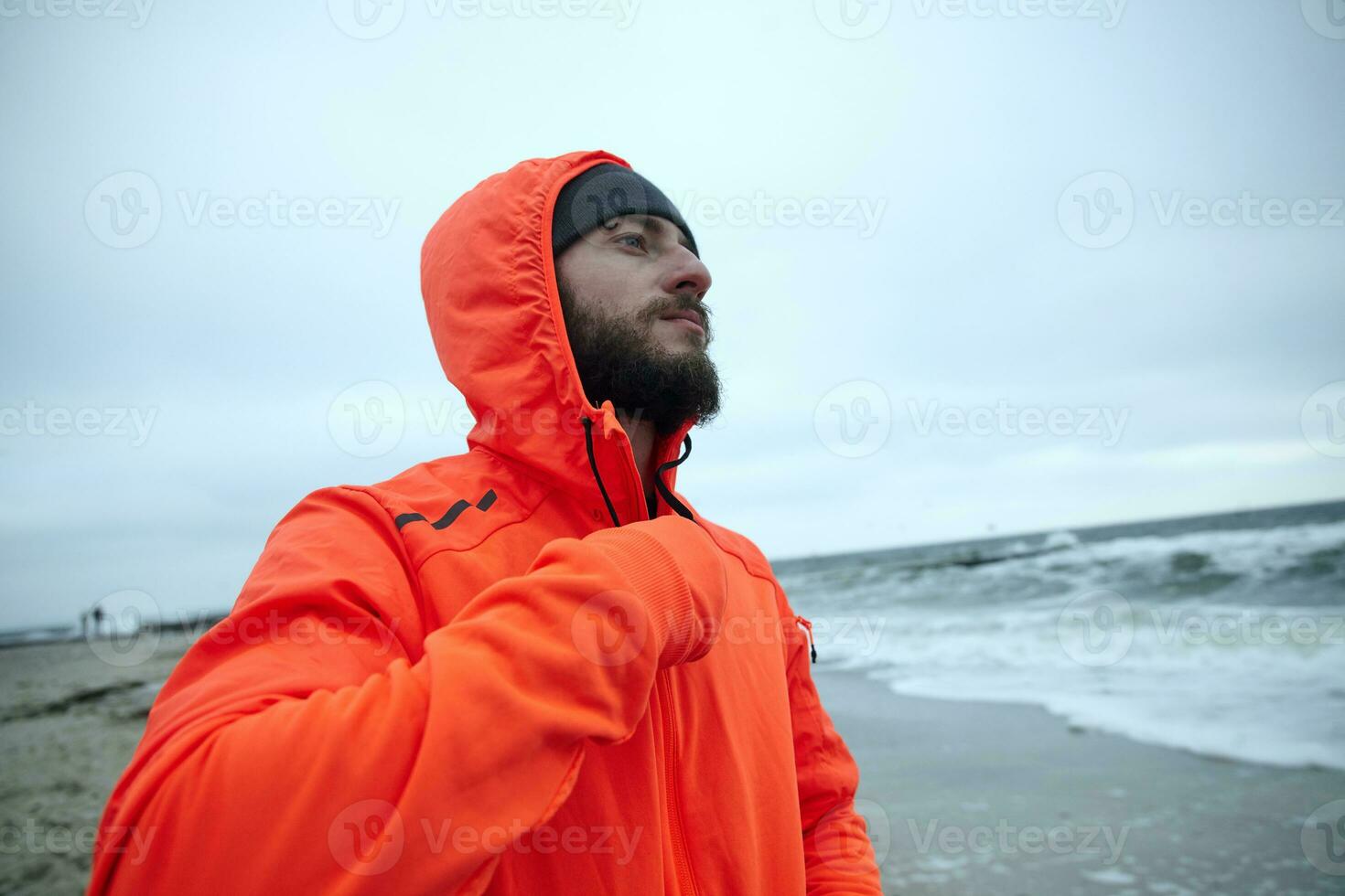 Shot of young sporty brunette bearded male standing over seaside on grey early morning after running session, wearing warm orange hoodie and black cap photo