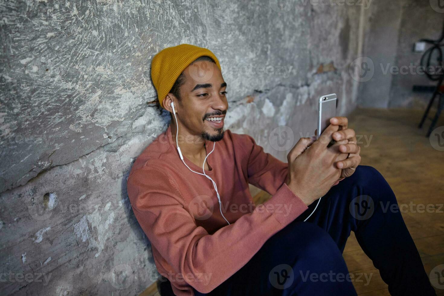 Positive pretty bearded dark skinned guy leaning on concrete wall while sitting on floor, wearing earphones and holding smartphone in raised hands, smiling while looking on screen photo