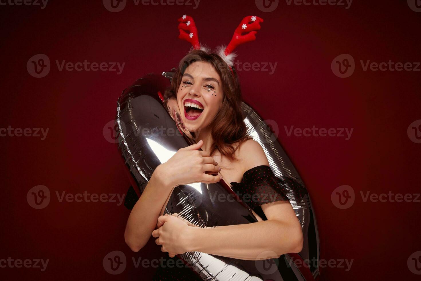 Studio shot of cheerful attractive young brown haired woman with evening makeup standing into number air balloon and laughing happily, wearing christmas horns while posing over claret background photo