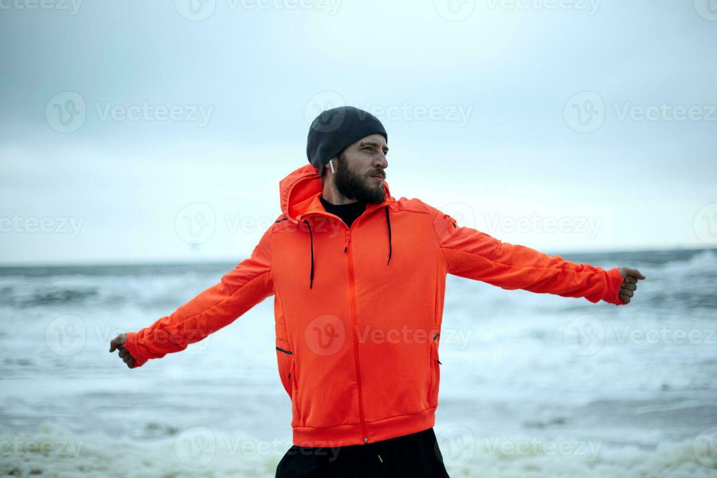 Outdoor shot of young dark haired sportsman with beard spreading his arms widely and breathing fresh air while walking over sandy beach after morning running session photo