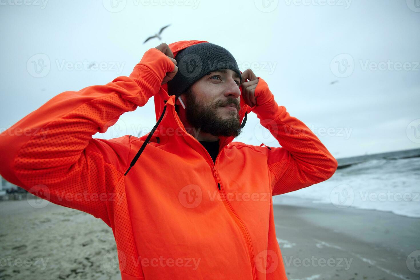 Positive young brunette bearded male in sporty clothes listening to music in his headphones and enjoying fresh weather over seaside, being calm and smiling slightly photo