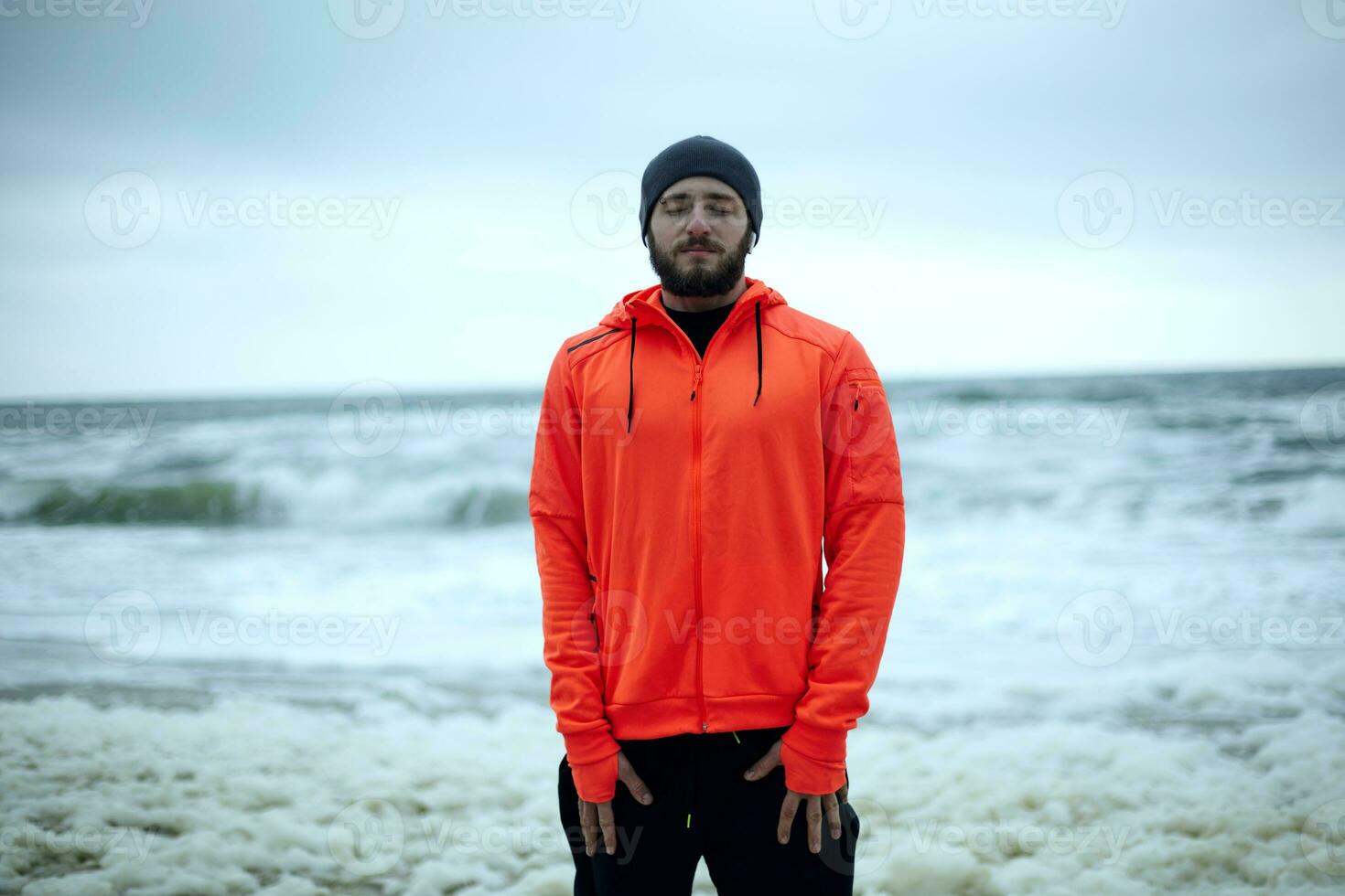 Image of young relaxed brunette bearded sportsman dressed in warm athletic clothes breathing deeply sea air with closed eyes after hard morning training over seaside photo