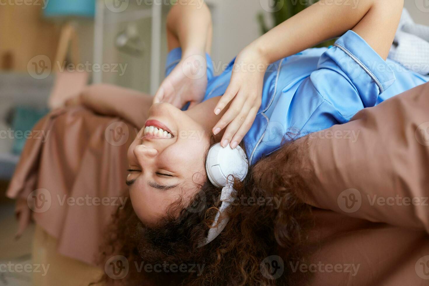 retrato de joven contento Rizado mulato niña acostado en el cama con su cabeza abajo, escuchando favorito música en auriculares, en general sonriente y mira disfrutando. foto
