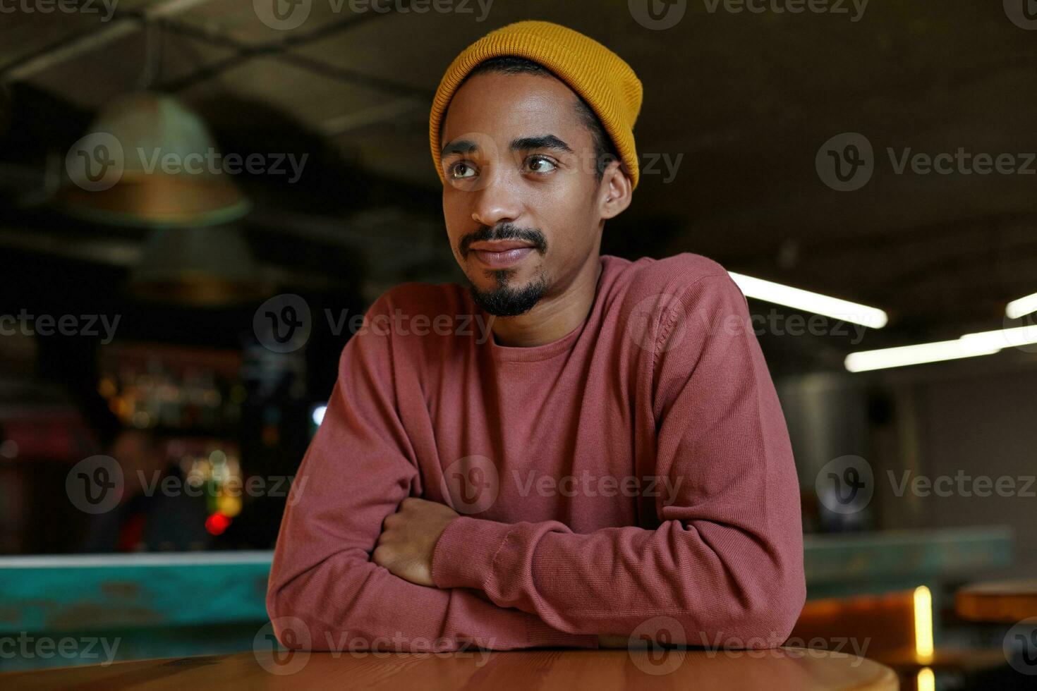 Portrait of pretty young bearded dark skinned male with charming brown eyes sitting at table and folding hands on it, looking aside with calm face and thinking about plans for evening photo