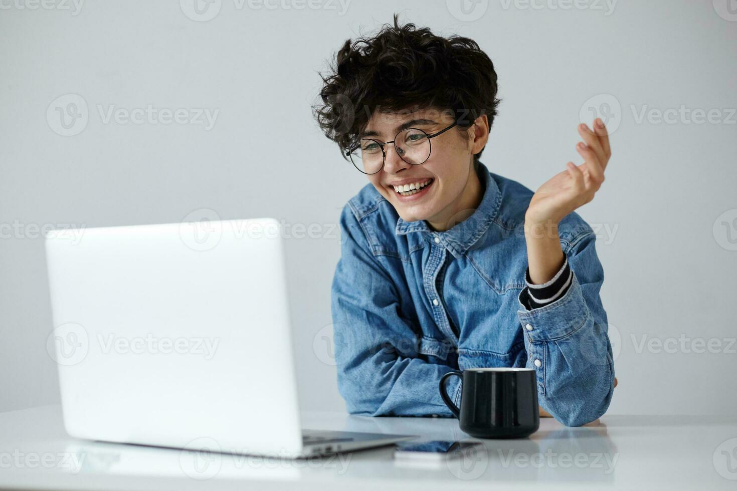 Cheerful young lovely curly brunette woman with short trendy haircut sitting over white background with modern laptop and having video call, smiling widely with raised palm photo