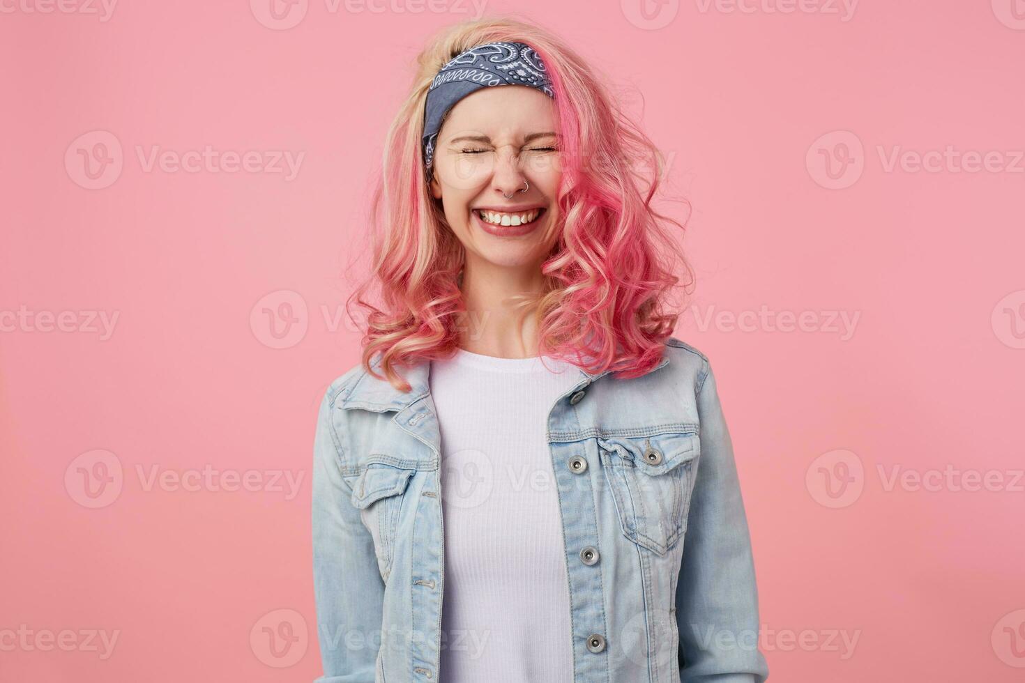 Happy cute smiling lady with pink hair and tattooed hands, standing over pink background with closed eyes, broadly smiling, wearing a white t-shirt and denim jacket. photo