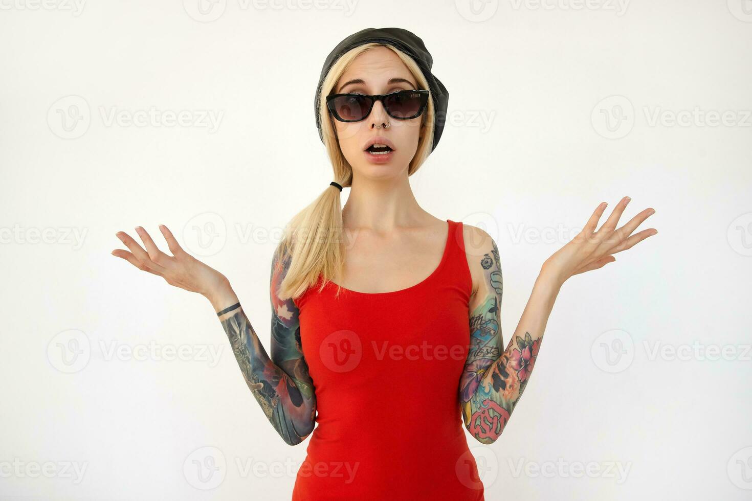 Puzzled young pretty blonde long haired woman with tattoos raising confusedly her hands while looking at camera, dressed in elegant clothes while posing over white background photo