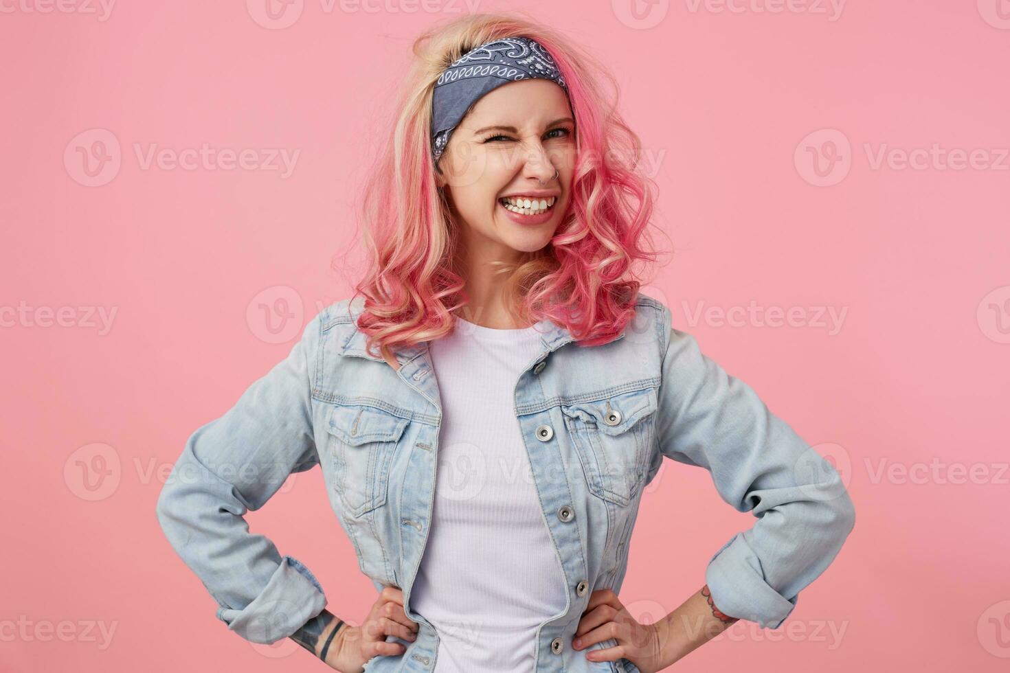 Portrait of happy cute girl with pink hair and tattooed hands, looking at the caera and winking, smiling and standing over pink background, wearing a white t-shirt and denim jacket. photo