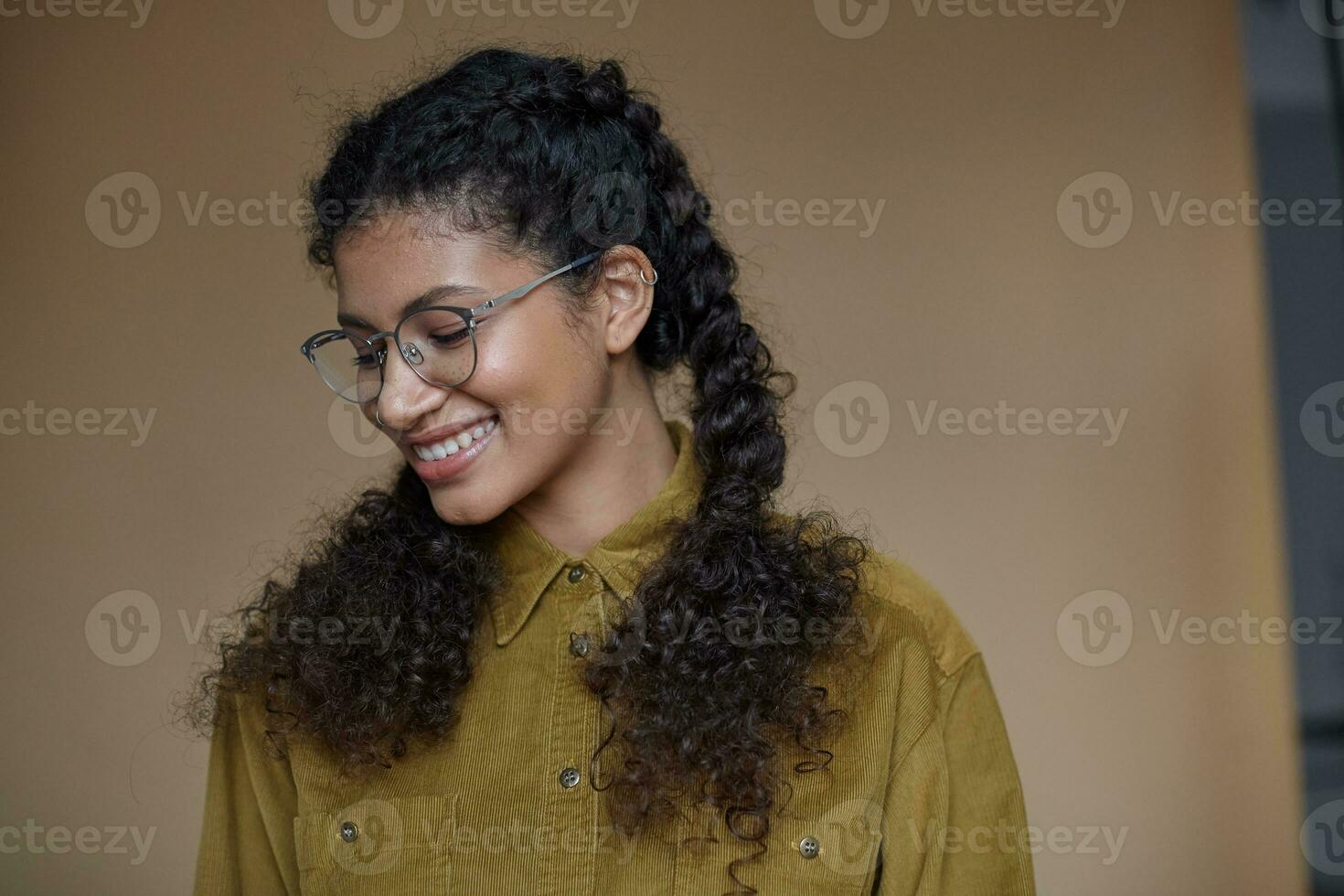 Indoor shot of good looking positive young dark skinned woman in glasses wearing her brown curly hair braided, dressed in mustard shirt while posing over beige background photo