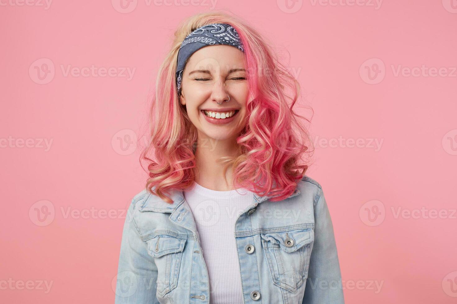 Happy cute smiling lady with pink hair and tattooed hands, waiting for surprise with closed eyes, broadly smiling, standing over pink background, wearing a white t-shirt and denim jacket. photo