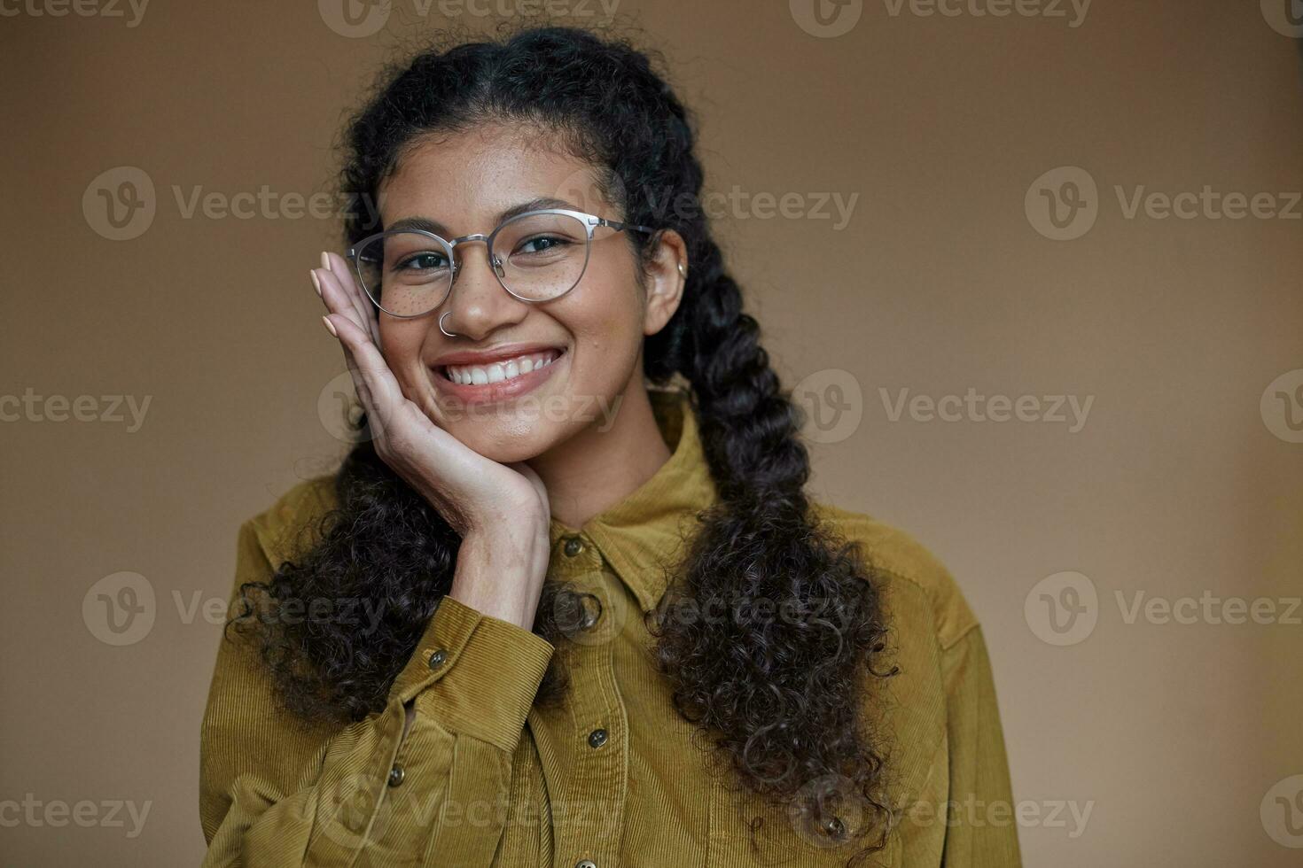 Portrait of cheerful pretty young dark skinned woman keeping her curly brown hair braided, looking happily to camera with wide smile and leaning chin on raised hand, isolated over beige background photo